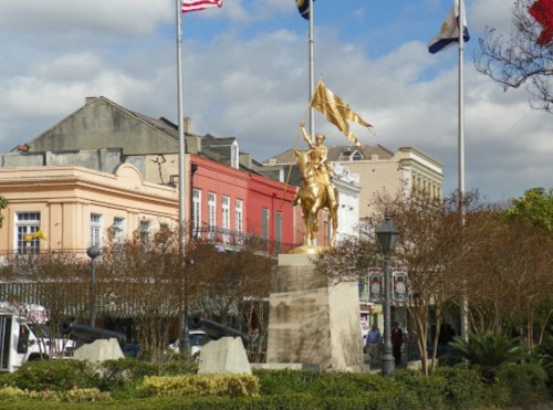 A golden statue of a mounted figure holding a flag, surrounded by trees and buildings, with flags flying above.