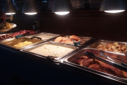 A buffet with various dishes, including meats, scrambled eggs, biscuits, and potatoes, under warm lighting.