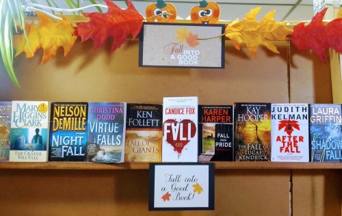 A bookshelf decorated with autumn leaves, featuring various books with fall-themed titles and a sign that says "Fall into a Good Book."
