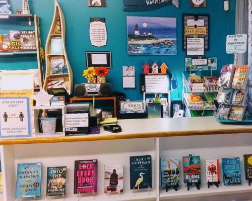 A cozy bookstore counter with books displayed, colorful decor, and a sign promoting social distancing.
