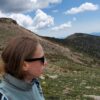 A person wearing sunglasses stands on a mountain, looking out at a scenic landscape with clouds in the sky.