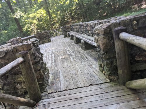 A wooden deck surrounded by stone walls and benches, set in a lush, green forest.