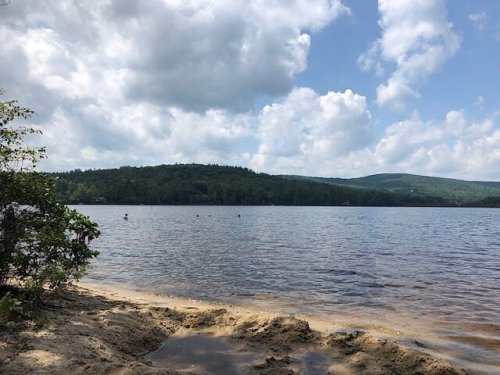 A serene lake scene with sandy shore, green hills, and fluffy clouds in a blue sky.
