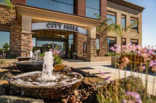 Modern city hall entrance with stone accents, water fountains, and landscaped flowers in front.