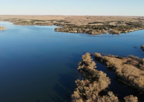 Aerial view of a calm lake surrounded by trees and rolling hills under a clear blue sky.