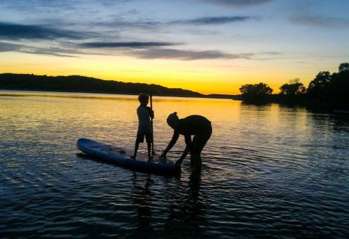 A silhouette of a child and an adult on a paddleboard at sunset, with calm water and a colorful sky in the background.