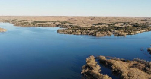 Aerial view of a serene lake surrounded by rolling hills and trees under a clear blue sky.