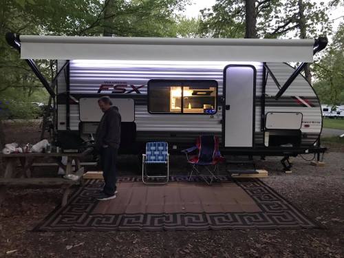 A camper trailer at a campsite with a man standing nearby, chairs set up, and a lighted awning.