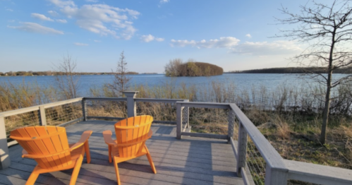 Two orange chairs on a wooden deck overlooking a calm lake with trees and a blue sky in the background.