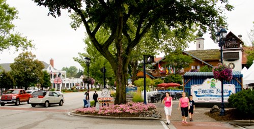 A street scene with shops, flowers, and people walking, set in a quaint town with a cloudy sky.