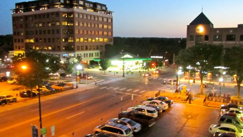 City street at dusk, featuring buildings, streetlights, and parked cars, with a gas station in the background.