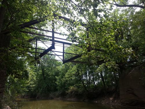 An old, rusted metal bridge frame partially obscured by lush green trees over a calm, reflective waterway.