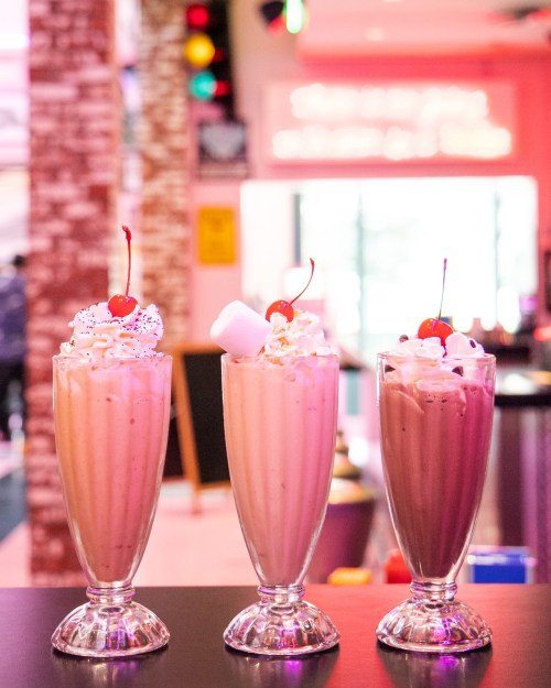 Three tall milkshakes in glasses, topped with whipped cream and cherries, displayed on a counter in a vibrant setting.