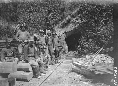 A group of miners poses near a mine entrance, with wooden beams and rocks scattered nearby.