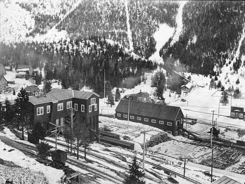 Historic black-and-white photo of a snowy mountain town with wooden buildings and surrounding trees.