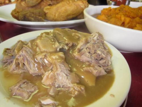 A plate of tender, shredded meat in gravy, served alongside fried chicken and a bowl of sweet potatoes.