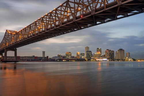 A bridge spans a river with a city skyline illuminated in the background under a cloudy sky at dusk.