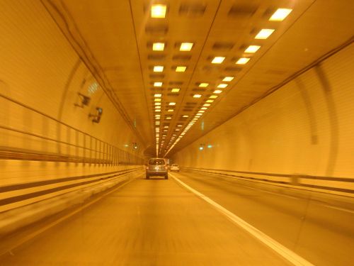 A car drives through a brightly lit tunnel with yellow walls and ceiling lights, creating a warm, reflective atmosphere.