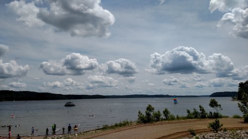 A serene lake scene with people enjoying the water, surrounded by lush greenery and a cloudy sky.