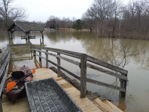 A wooden dock leads to a flooded area with a kayak and a boat visible, surrounded by trees and cloudy skies.