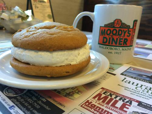 A cream-filled cookie sandwich on a plate next to a coffee mug from Moody's Diner in Waldoboro, Maine.