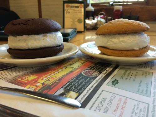 Two whoopie pies on plates: one chocolate with white filling, one vanilla with white filling, on a table with a menu.