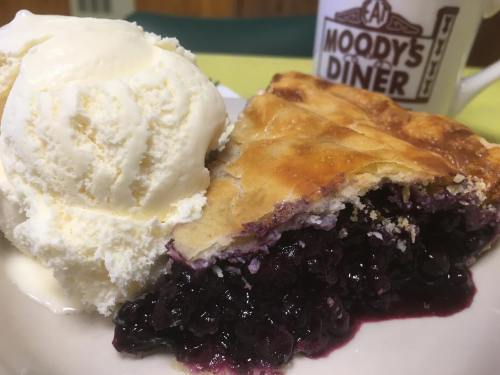 A slice of blueberry pie with a scoop of vanilla ice cream on a plate, next to a mug from Moody's Diner.