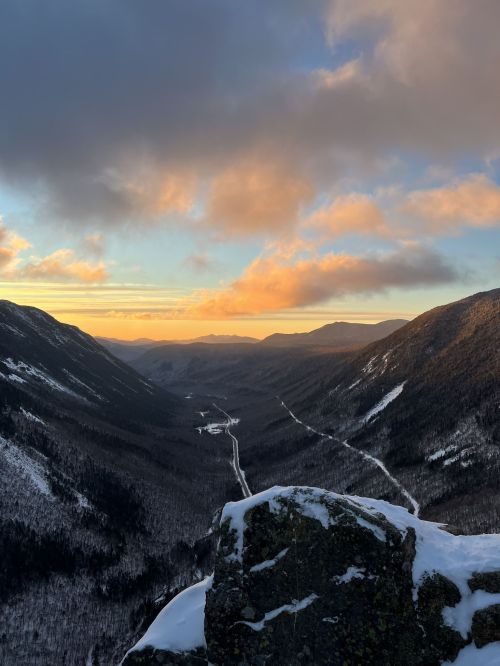 A scenic view of a snow-covered valley at sunset, with mountains and a winding road visible in the distance.