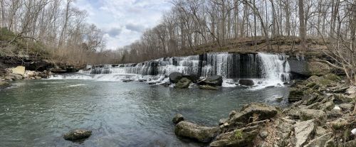 A serene waterfall cascades over rocks into a calm river, surrounded by bare trees and a cloudy sky.