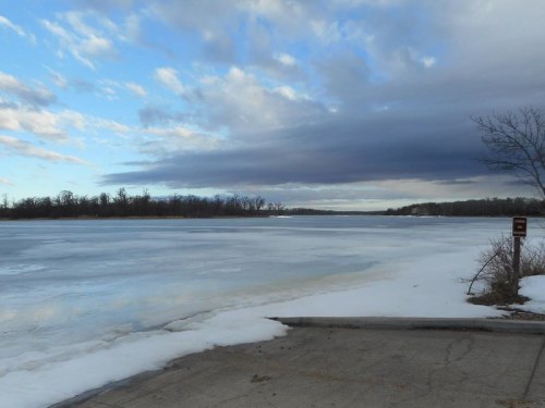 A frozen lake under a cloudy sky, with patches of ice and snow along the shore.