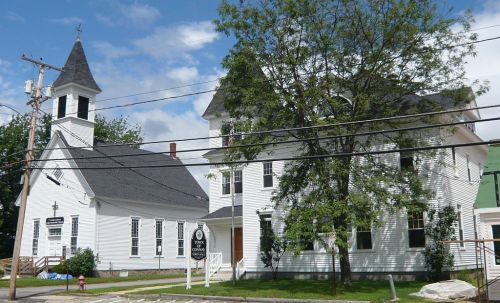 A white church building with a steeple, surrounded by trees and power lines, under a partly cloudy sky.