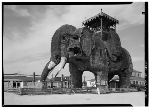 A large wooden elephant structure with a tower on its back, surrounded by a fence and buildings in the background.