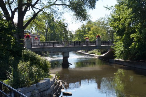 A serene view of a bridge over a calm river, surrounded by lush greenery and flower planters.