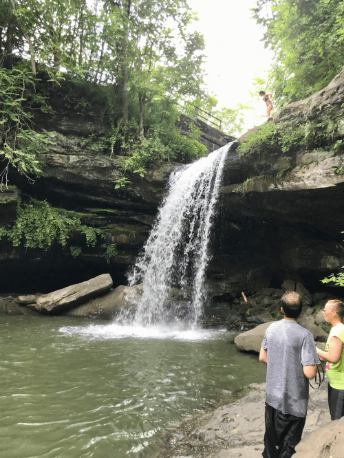 A waterfall cascades over rocks into a serene pool, surrounded by lush greenery and people enjoying the scene.