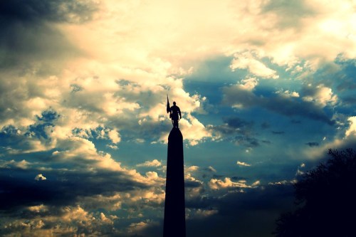 Silhouette of a statue atop a tall column against a dramatic sky filled with clouds.