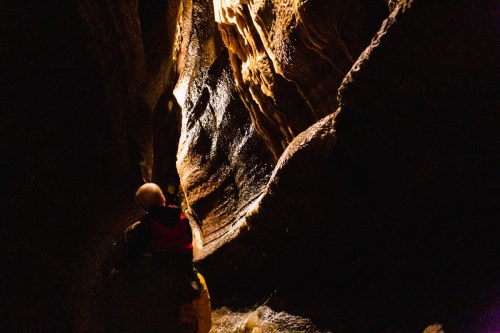 A person in a kayak explores a dark cave with illuminated rock formations and water flowing nearby.