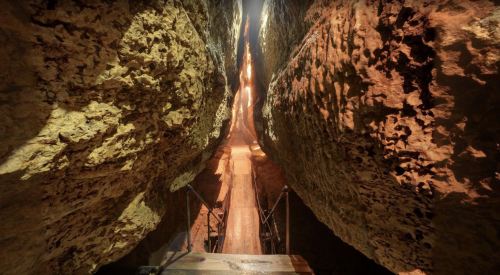 A narrow canyon with steep rock walls and a wooden bridge leading through the crevice, illuminated by soft light.