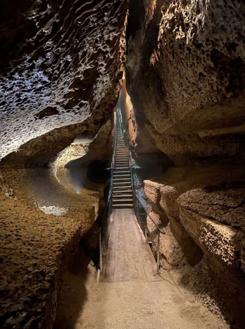 A narrow cave passage with textured rock walls and wooden stairs leading upward in dim lighting.