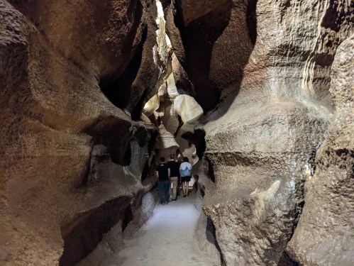 Two people walking through a narrow, textured cave passage with rocky walls and soft lighting.