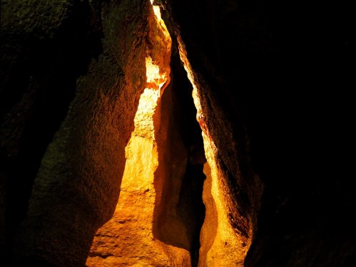 A narrow crevice in a dark cave illuminated by warm, golden light, highlighting the textured rock formations.