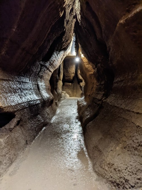 A narrow, dimly lit cave passage with smooth, wet walls and a sandy floor, leading into darkness.