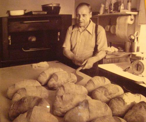 A baker in suspenders slices freshly baked loaves of bread on a countertop, with an oven in the background.