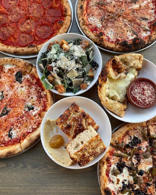 A spread of various pizzas, a Caesar salad, and breadsticks with dipping sauce on a wooden table.