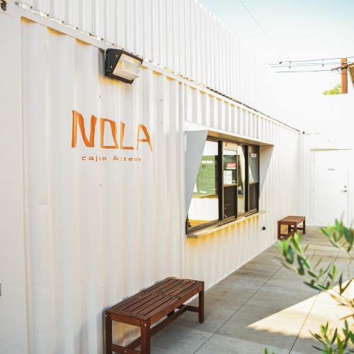Exterior of a container-style building with "NOLA cajun & creole" sign, featuring a window and wooden benches.