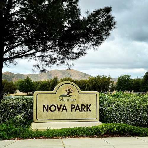 Sign for Nova Park in Menifee, surrounded by greenery and mountains under a cloudy sky.