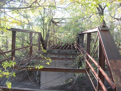 An old, rusty bridge surrounded by trees, with overgrown vegetation and a concrete base visible beneath.