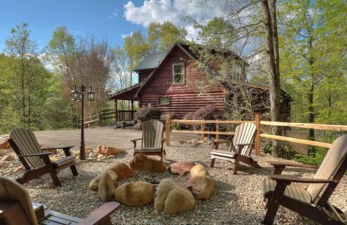 A rustic cabin surrounded by trees, with a fire pit and wooden chairs in the foreground on a sunny day.