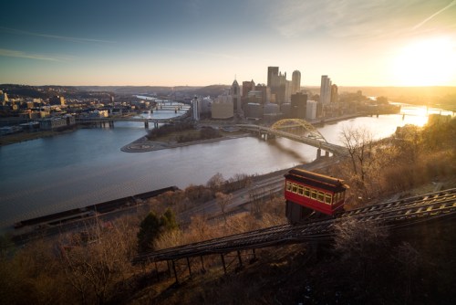 A scenic view of Pittsburgh at sunset, featuring a funicular railway and rivers with city skyline in the background.