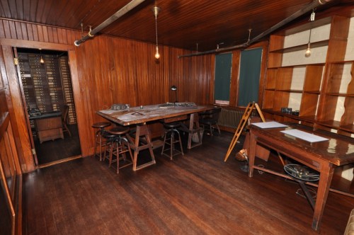 A wooden-paneled room with a large table, chairs, and shelves, featuring warm lighting and a vintage atmosphere.