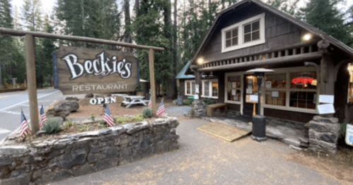 A rustic restaurant with a wooden sign reading "Beckie's Restaurant," surrounded by trees and flags, inviting visitors.